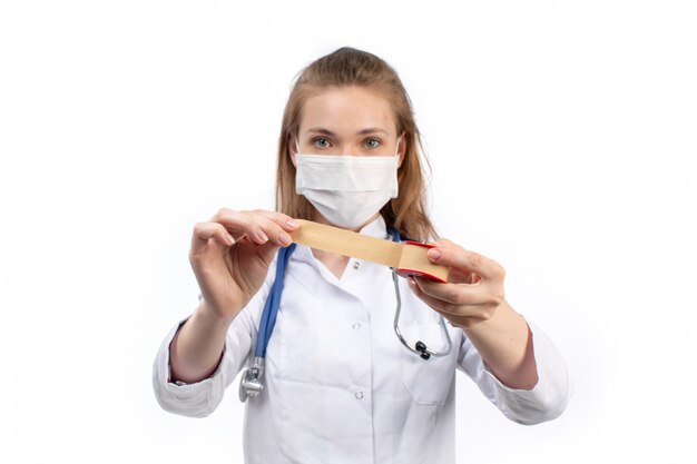 Joven doctora en traje médico blanco con máscara protectora blanca posando sosteniendo vendaje médico en el blanco