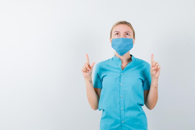 Joven doctora en uniforme apuntando hacia arriba y mirando confiado