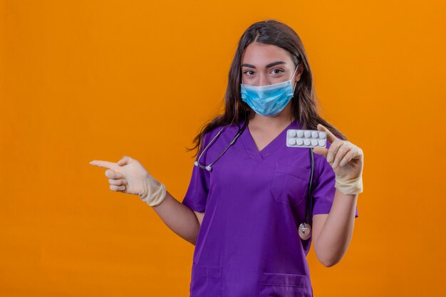 Joven doctora en uniforme médico con fonendoscopio con máscara protectora y guantes con ampolla con píldoras y apuntando con el dedo hacia un lado sobre fondo naranja aislado