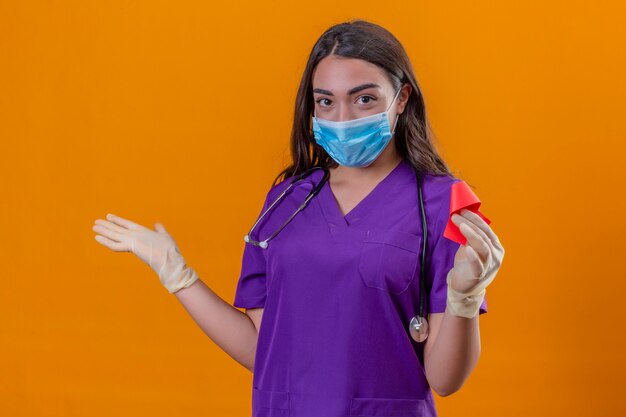 Joven doctora en uniforme médico con fonendoscopio con máscara protectora y guantes sonriendo sosteniendo la cinta roja de pie con el brazo en un gesto de bienvenida sobre fondo naranja aislado