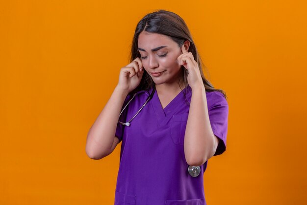 Joven doctora en uniforme médico con fonendoscopio de pie con los ojos cerrados sensación escalonada con fuerte dolor de cabeza sobre fondo naranja aislado