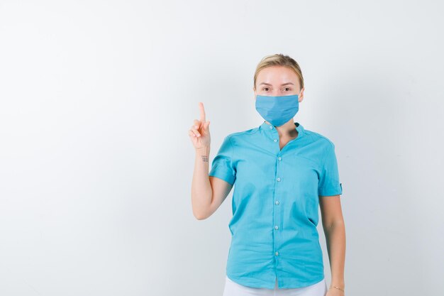 Joven doctora en uniforme médico, máscara apuntando hacia arriba y mirando confiada