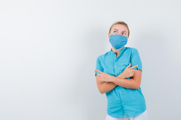 Joven doctora en uniforme médico, máscara apuntando a derecha e izquierda y mirando vacilante