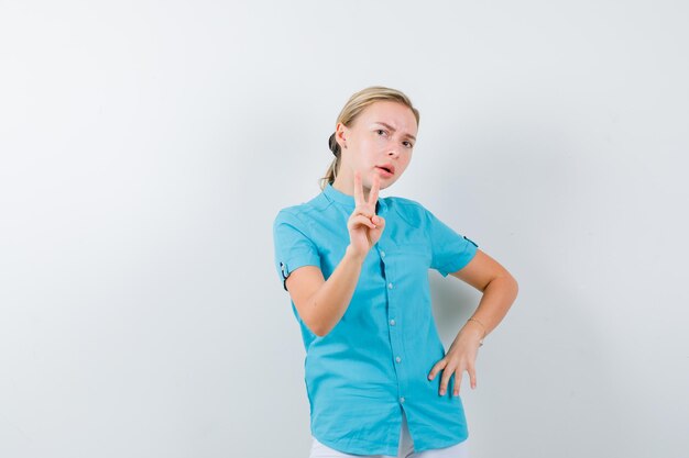 Joven doctora en uniforme médico, máscara mostrando el signo de la victoria y mirando desconcertado