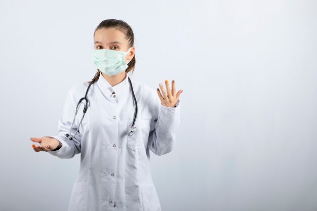 Joven doctora en uniforme médico y máscara de pie sobre fondo blanco.