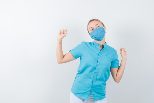 Joven doctora en uniforme médico, máscara que muestra el gesto del ganador y se ve feliz