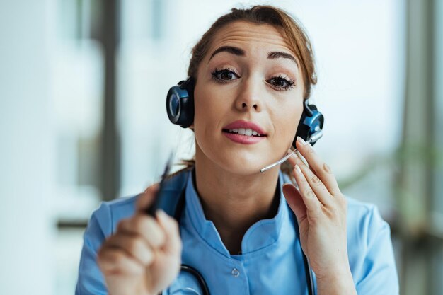 Joven doctora usando auriculares y hablando con pacientes mientras trabaja en el centro de llamadas médicas y mira la cámara