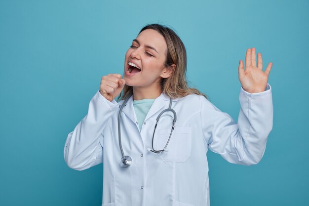 Joven doctora vistiendo bata médica y estetoscopio alrededor del cuello finge sostener el micrófono manteniendo la mano en el aire cantando con los ojos cerrados