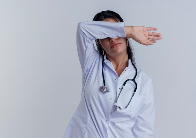 Joven doctora vistiendo bata médica y estetoscopio cubriendo los ojos con el brazo aislado