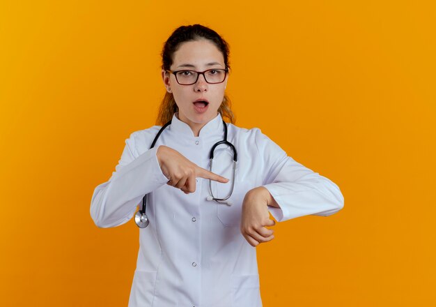 Joven doctora vistiendo bata médica y estetoscopio con gafas mostrando gesto de reloj de pulsera aislado