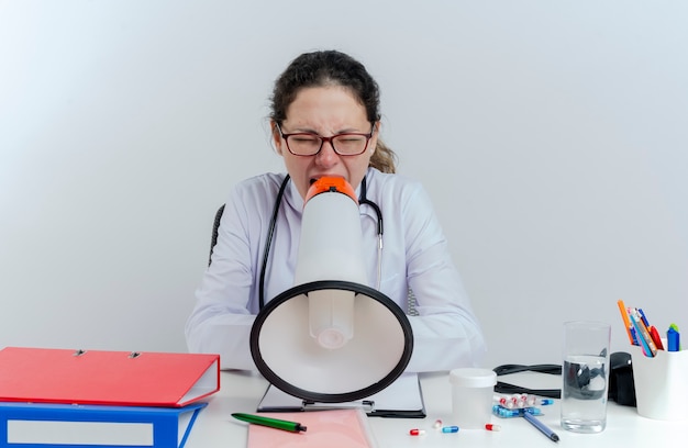 Joven doctora vistiendo bata médica y estetoscopio y gafas sentado en el escritorio con herramientas médicas gritando en altavoz con los ojos cerrados aislados