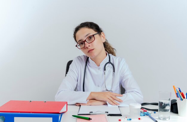 Joven doctora vistiendo bata médica y estetoscopio y gafas sentado en el escritorio con herramientas médicas poniendo las manos sobre el escritorio mirando aislado