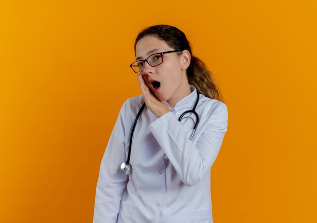 Joven doctora vistiendo bata médica y estetoscopio con gafas susurra aislado