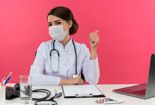 Joven doctora vistiendo bata médica con estetoscopio en máscara médica sentado en el escritorio trabajar en la computadora con herramientas médicas apunta al lado de la pared rosa con espacio de copia
