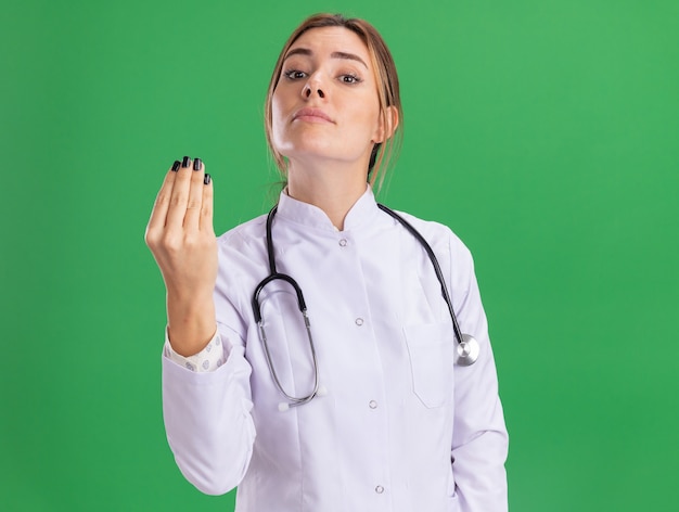 Joven doctora vistiendo bata médica con estetoscopio mostrando el gesto de la punta aislado en la pared verde