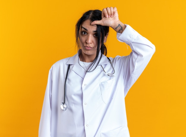 Joven doctora vistiendo bata médica con estetoscopio mostrando el pulgar hacia abajo aislado en la pared amarilla