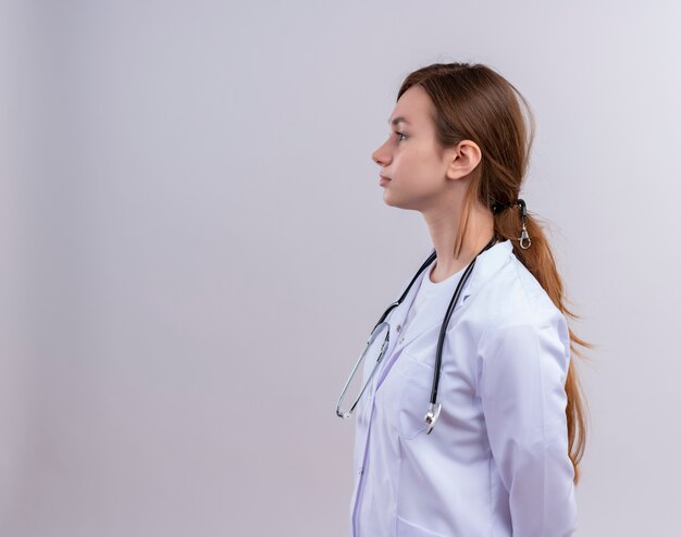 Joven doctora vistiendo bata médica y estetoscopio de pie en la vista de perfil en la pared blanca aislada con espacio de copia