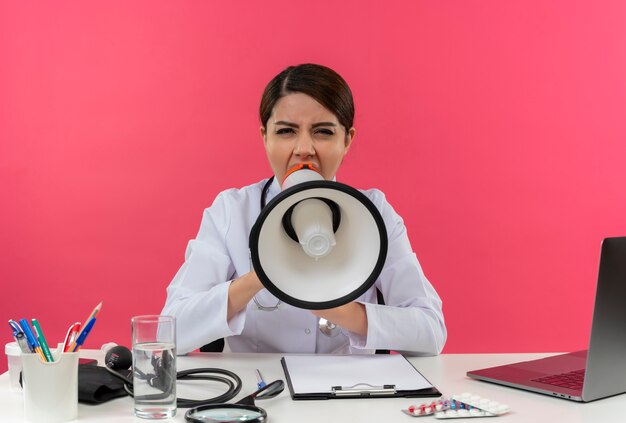 Joven doctora vistiendo bata médica con estetoscopio sentado en el escritorio, trabajo en computadora con herramientas médicas, habla por altavoz en una pared rosa aislada con espacio de copia