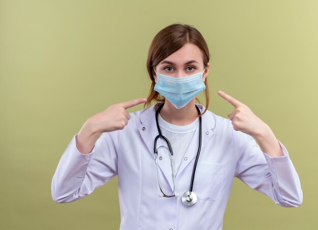 Joven doctora vistiendo bata médica, máscara y estetoscopio apuntando a su máscara en la pared verde aislada