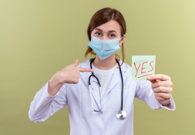 Joven doctora vistiendo bata médica, máscara y estetoscopio estirando la nota de sí y apuntando a sí misma en la pared verde aislada