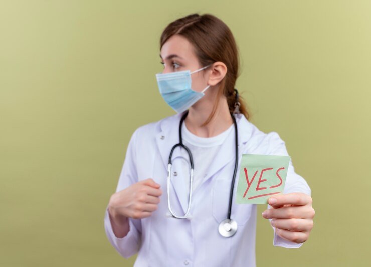 Foto gratuito joven doctora vistiendo bata médica, máscara y estetoscopio estirando sí nota mirando el lado izquierdo en la pared verde aislada con espacio de copia