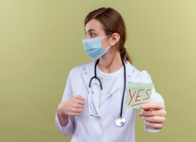 Joven doctora vistiendo bata médica, máscara y estetoscopio estirando sí nota mirando el lado izquierdo en la pared verde aislada con espacio de copia