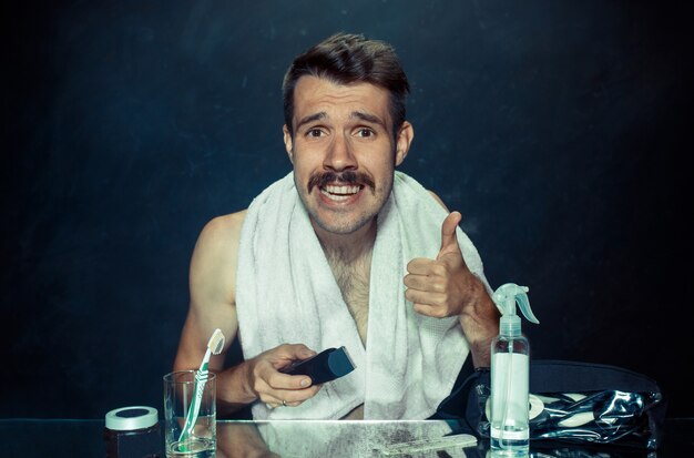 El joven en el dormitorio sentado frente al espejo rascándose la barba en su casa. Concepto de emociones humanas