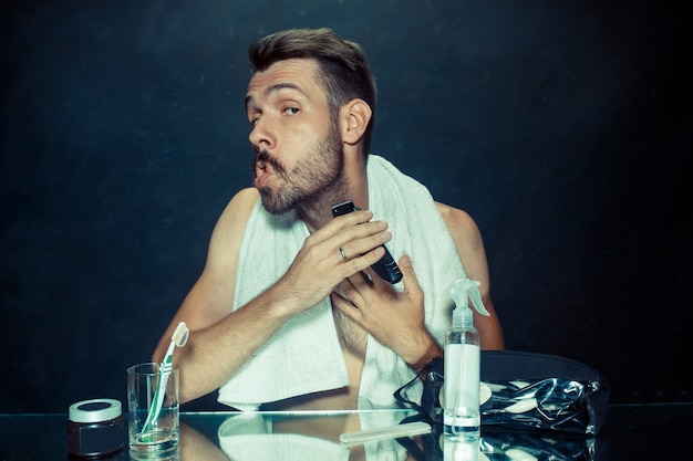 El joven en el dormitorio sentado frente al espejo rascándose la barba en su casa. Concepto de emociones humanas