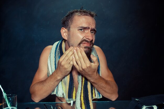 El joven en el dormitorio sentado frente al espejo rascándose la barba en su casa. Concepto de emociones humanas