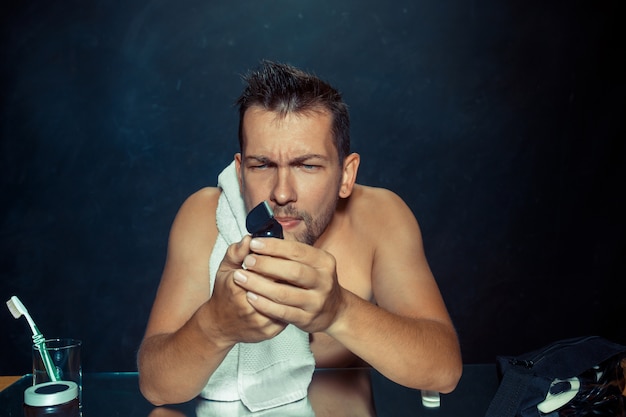 El joven en el dormitorio sentado frente al espejo rascándose la barba en su casa. Concepto de emociones humanas