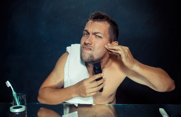 El joven en el dormitorio sentado frente al espejo rascándose la barba en su casa. Concepto de emociones humanas