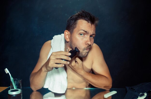 El joven en el dormitorio sentado frente al espejo rascándose la barba en su casa. Concepto de emociones humanas