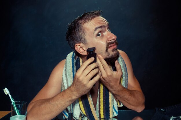 El joven en el dormitorio sentado frente al espejo rascándose la barba en su casa. Las emociones humanas y el concepto de estilo de vida.