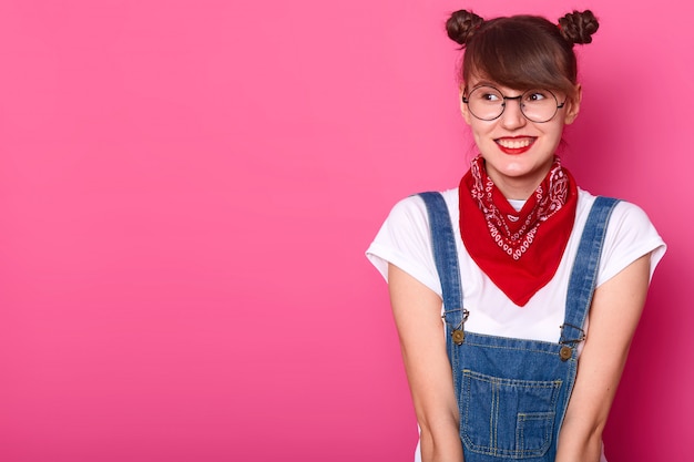 Joven dulce niña sonriente mirando a un lado, vistiendo camiseta blanca casual, overol de jeans, pañuelo rojo y gafas redondas de moda, estar de buen humor, tener racimos. Copiar espacio para publicidad.