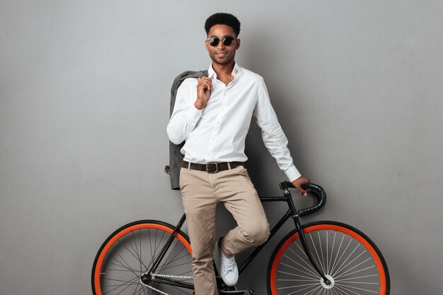 Joven elegante afroamericano de pie y apoyado en bicicleta