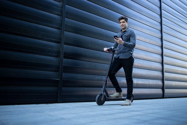 Joven elegante caminando con scooter eléctrico y usando teléfonos inteligentes.