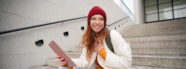 Foto gratuita joven y elegante estudiante pelirroja sentada en las escaleras al aire libre con tableta digital lee usos