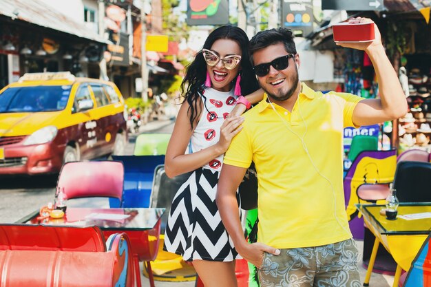 Joven elegante hipster hermosa pareja sentada en la colorida cafetería, coqueta, traje de moda, traje de moda, gafas de sol, vacaciones tropicales, romance de vacaciones, luna de miel, sonriendo, feliz, escuchando música