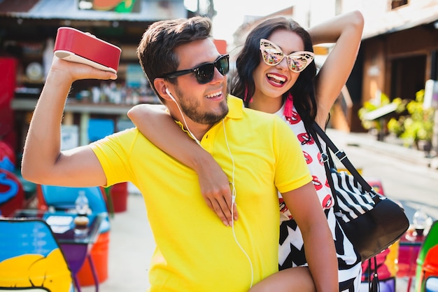 Joven elegante hipster hermosa pareja sentada en la colorida cafetería, coqueta, traje de moda, traje de moda, gafas de sol, vacaciones tropicales, romance de vacaciones, luna de miel, sonriendo, feliz, escuchando música