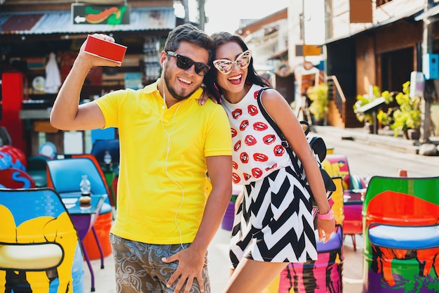 Joven elegante hipster hermosa pareja sentada en la colorida cafetería, coqueta, traje de moda, traje de moda, gafas de sol, vacaciones tropicales, romance de vacaciones, luna de miel, sonriendo, feliz, escuchando música