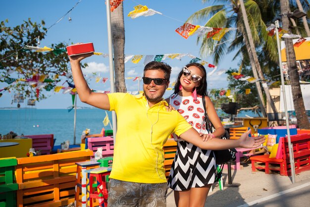 Joven elegante hipster hermosa pareja en vacaciones de verano en Tailandia, coqueta, atuendo de moda, gafas de sol, romance tropical, sonriente, feliz, escuchando música, humor de fiesta, café en la playa