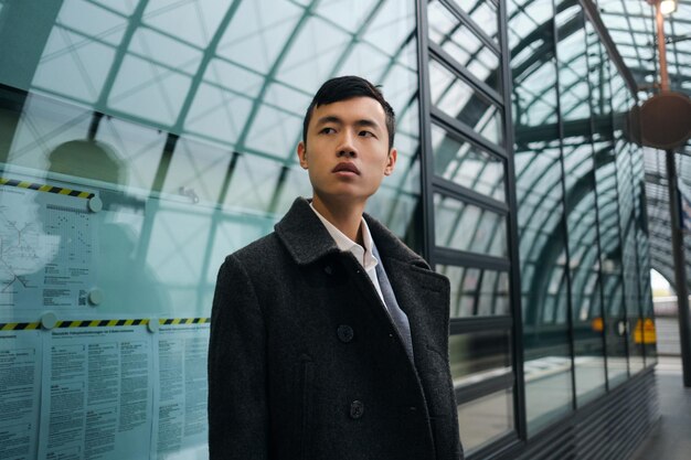 Joven y elegante hombre de negocios asiático con abrigo mirando atentamente hacia otro lado esperando el tren en la moderna estación de metro
