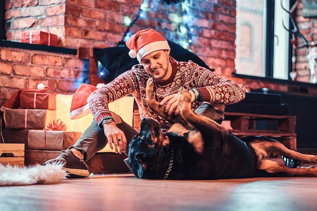 Un joven elegante jugando con su lindo perro en una sala de estar decorada en Navidad.
