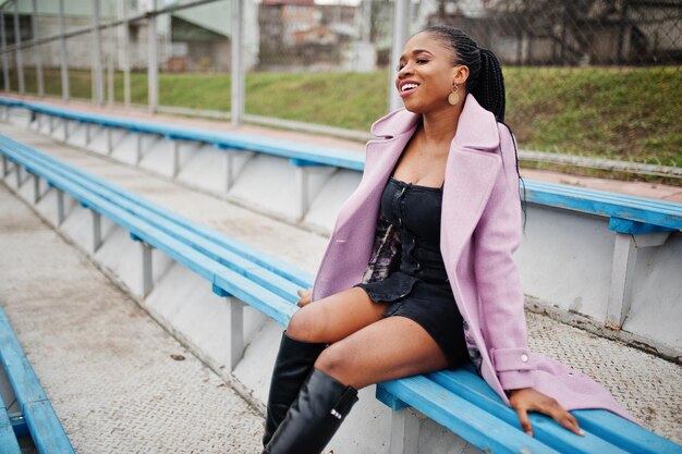Joven y elegante mujer afroamericana en la calle en las gradas del estadio con abrigo de moda