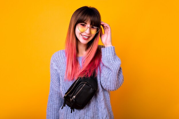 Joven y elegante mujer inconformista maravillosa con largos pelos fucsia ombre posando en la pared amarilla, vibraciones primaverales, colores pastel suaves, gafas de sol de corazón vintage y riñonera de moda.