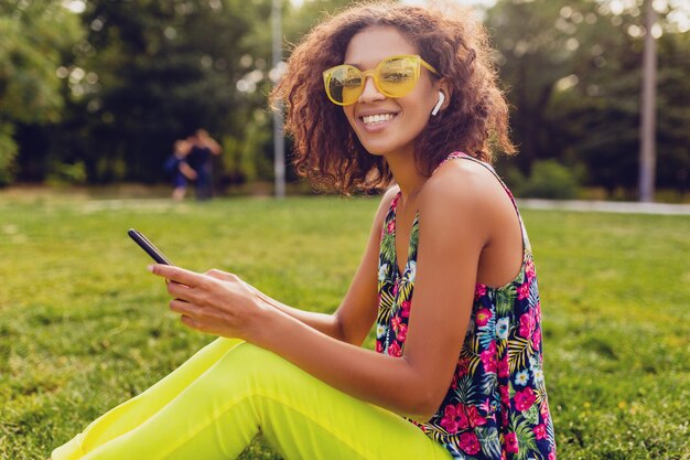 Joven y elegante mujer negra sonriente con smartphone escuchando música con auriculares inalámbricos divirtiéndose en el parque, estilo colorido de la moda de verano, sentado en la hierba, gafas de sol amarillas