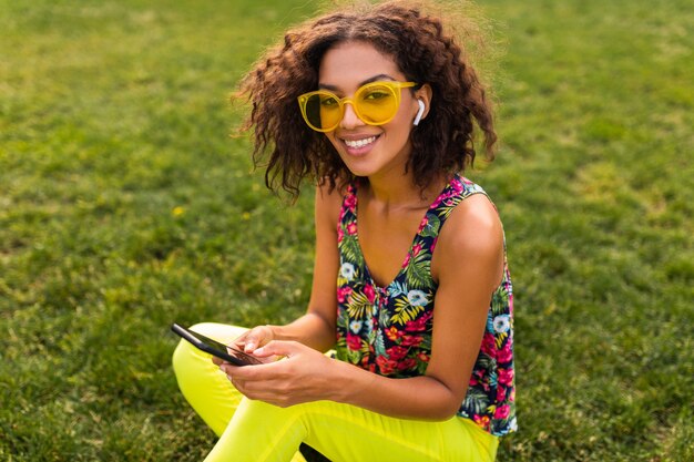 Joven y elegante mujer negra sonriente con smartphone escuchando música con auriculares inalámbricos divirtiéndose en el parque