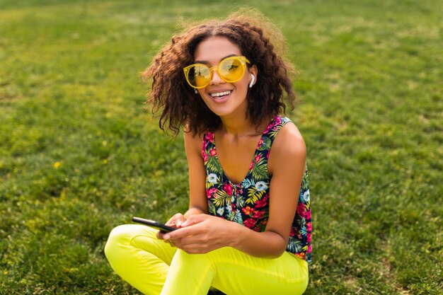 Joven y elegante mujer negra sonriente con smartphone escuchando música con auriculares inalámbricos divirtiéndose en el parque