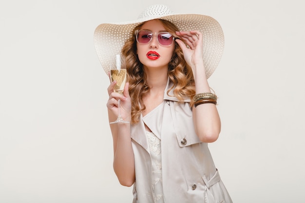 Joven y elegante mujer rubia sonriente con sombrero grande y gafas de sol con copa de champán en una fiesta feliz