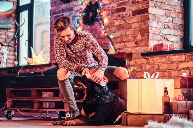 Un joven elegante que sostiene una caja de regalo mientras se sienta con su lindo perro en una sala de estar decorada en Navidad.